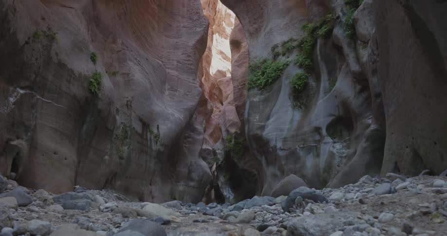 A small stream that comes out of a hole in the middle of tall rock formations and there is a lot of gravel around it