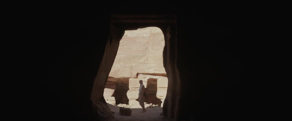 A woman walks to the entrance of a cave in the desert before walking off
