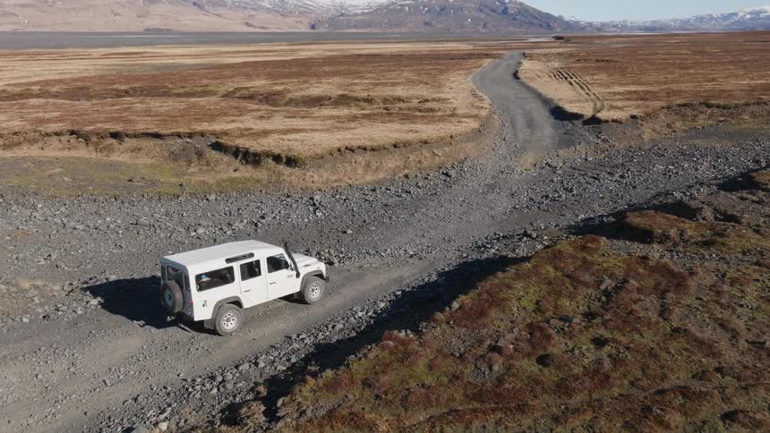 A white jeep stops on a dirt road amidst ruins and mountains on the horizon covered in snow