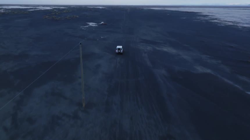 Aerial view of a white jeep driving on black sand beach near the frozen lake