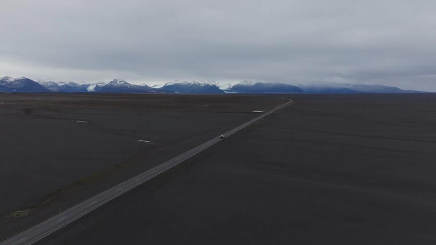 An aerial view of a very large flat terrain with mountains in the background and a road cutting across the landscape on an overcast day