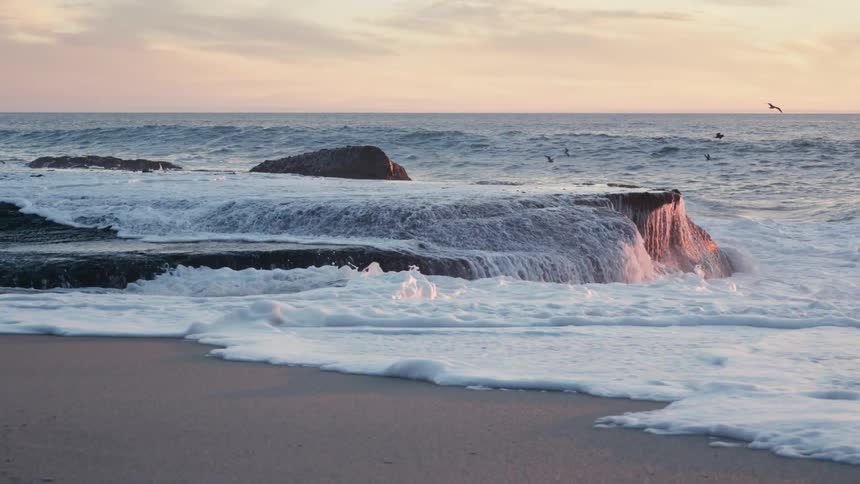 Ocean waves break against the beach rhythmically colliding with resilient rocks along the shore