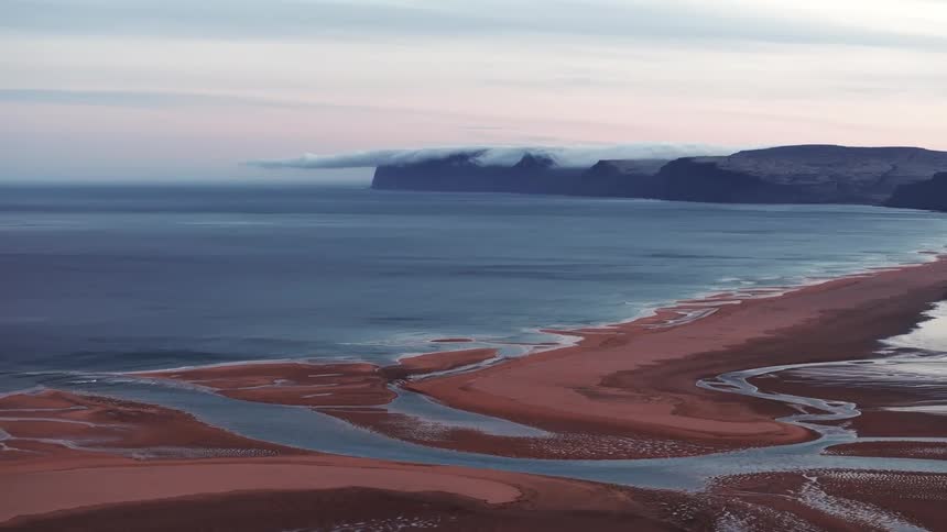 Aerial panorama of the sand in the middle of the sea with a mountain range on the horizon under a dawn sky