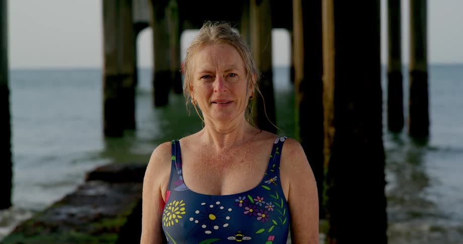 Older woman stands smiling underneath pier in a swimsuit