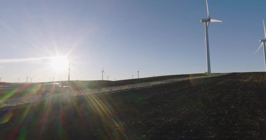 Car driving on a road next to a grassland with wind energy platforms on a day with a blue sky and sunlight reflection