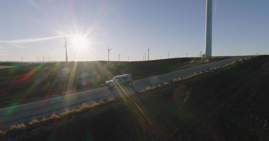 A truck travels on a rural road through a wind farm as the sun shines brightly in the distance