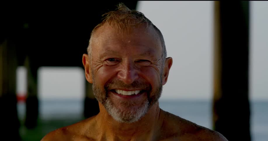 Beneath a pier a man stands smiling at the camera on the beach