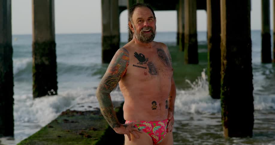 Older man wearing swimsuit stands under pier smiling as waves roll forward in the background