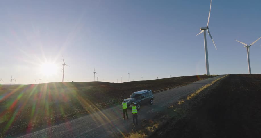 Sun glares onto two utility workers having a discussion near vehicle parked on road passing wind farm