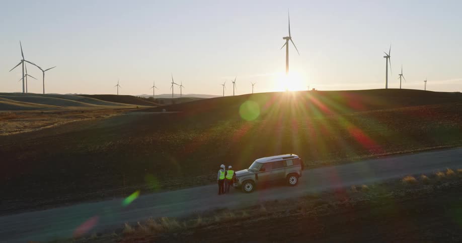 Two people in safety gear next to a car amidst fields with wind energy turbines on a morning