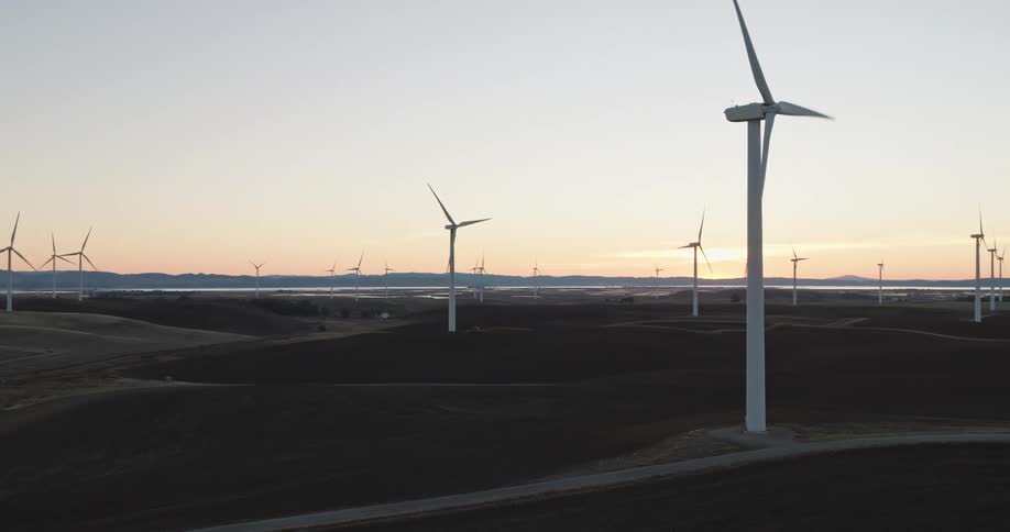 Multiple roads cut through wind farm bordering water under a red tinted sky on a foggy day