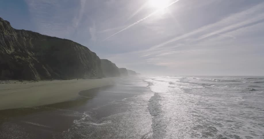 Waves roll into shore and over the beach near a promontory as a person walks on the beach