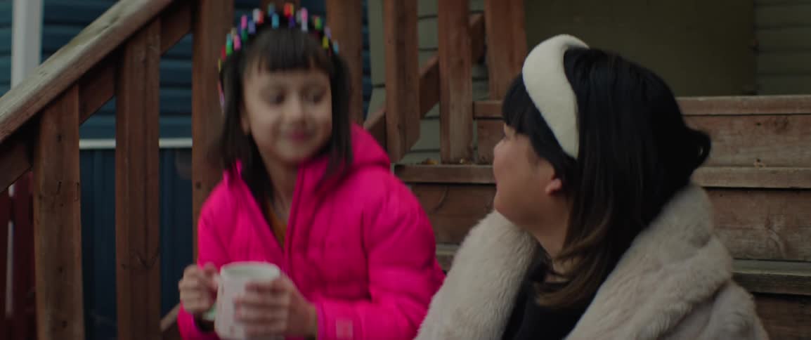 A woman is sitting outside on a porchs steps when a young girl drinking from a mug comes out and joins her