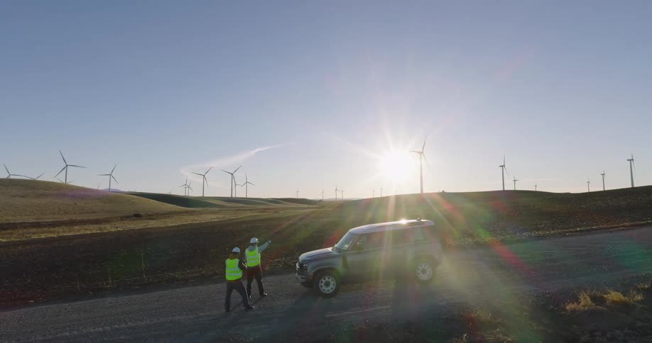 Utility workers discuss wind turbines while standing next to parked car on a sunny day