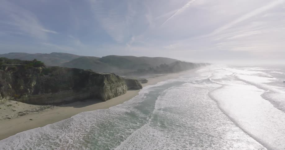 Waves roll into shore and over a beach at the foot of mountain cliffs along the coast