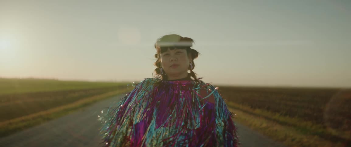 Woman stands in a rural road wearing an outfit covered in metallic multicolored strips at sunset