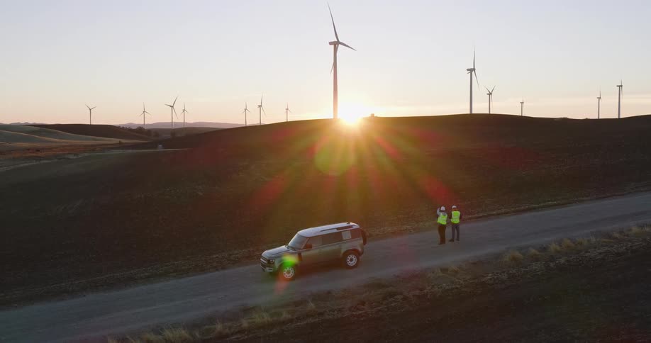 Utility workers stand near a truck on a road as wind turbines spin on the hills