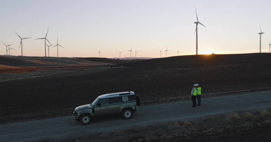 Workers stand on a access road collecting aerial footage of the turbines on a wind farm at sunset