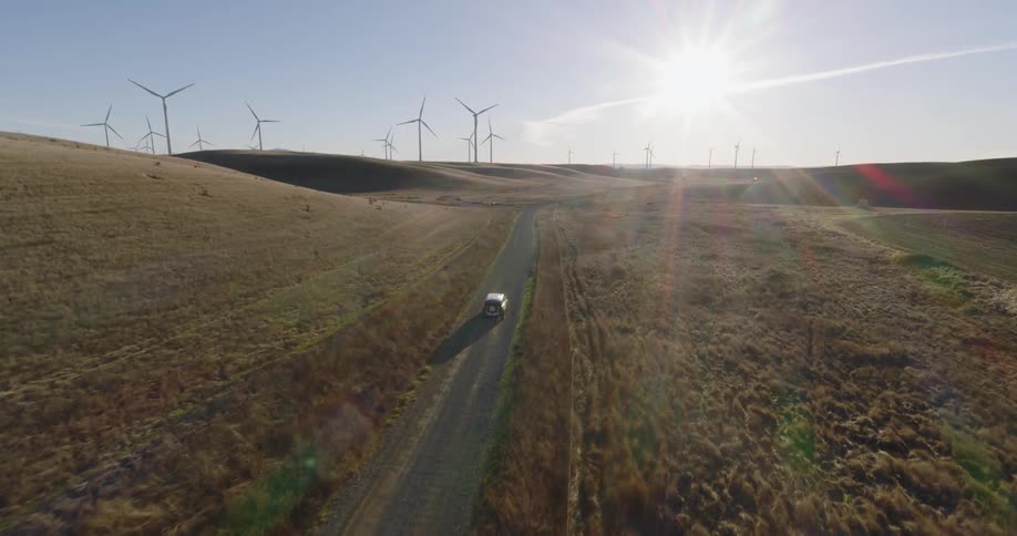 A truck travels on a rural road through a wind farm as the sun shines brightly in the distance