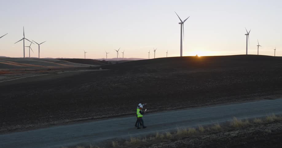 Utility workers walk away from a van on a road as wind turbines spin on the hills