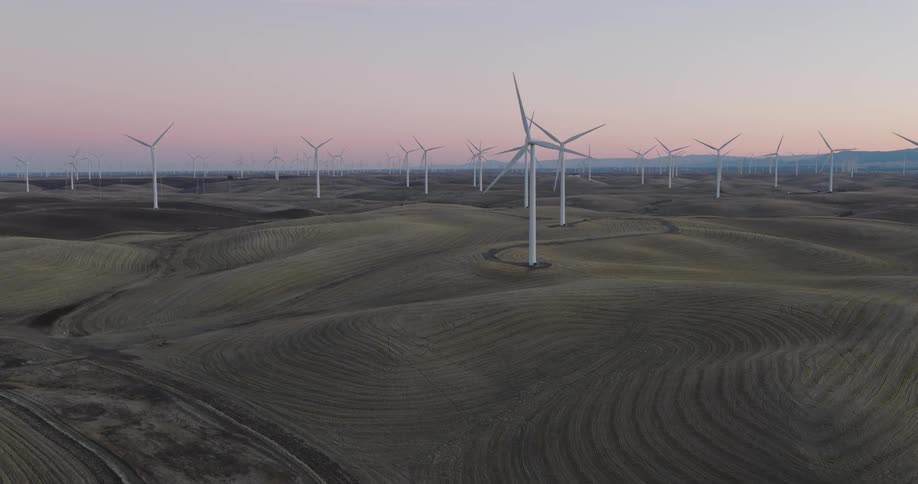 Wind turbines turning slowly against a red tinted horizon tower over hills with mowing lines