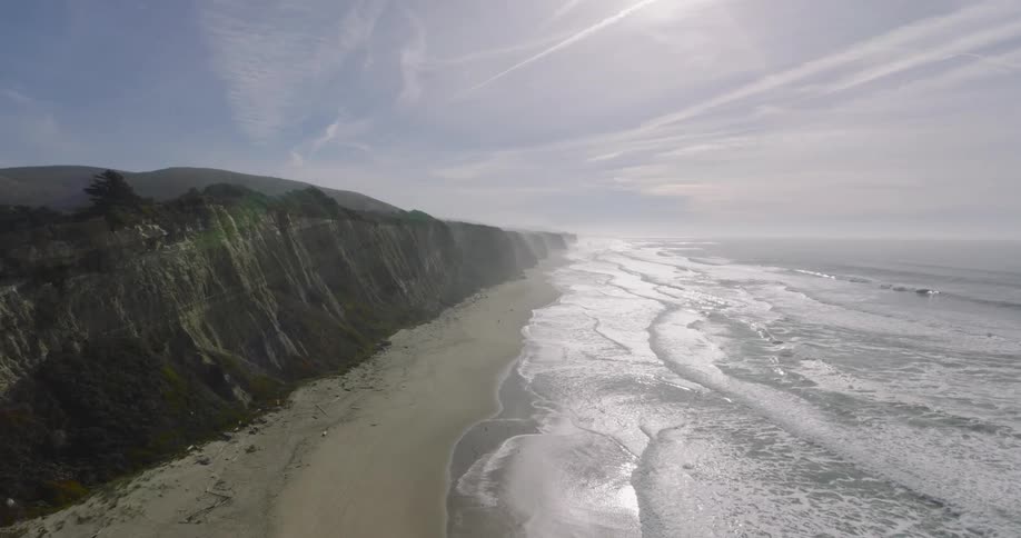 Aerial of waves crashing on a thin cliff lined beach with the sunrays glaring across the sky and reflecting from the water