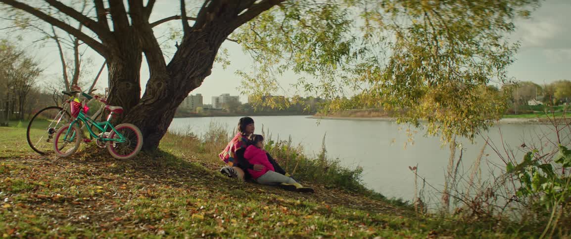 A mother and daughter sit by the river's edge in a park during sunset