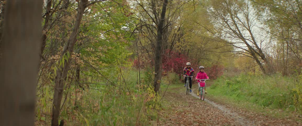 Mother and daughter pedal bicycles along trail through woods in autumn