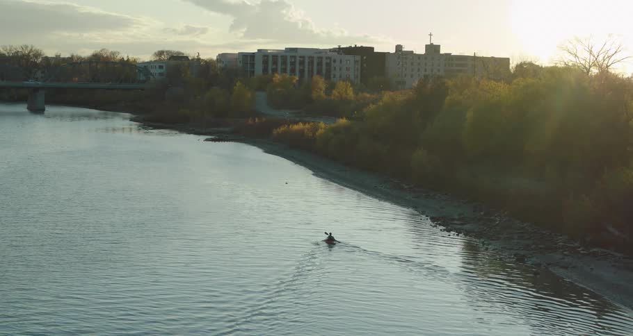 A person paddles a kayak down a city river at sunset