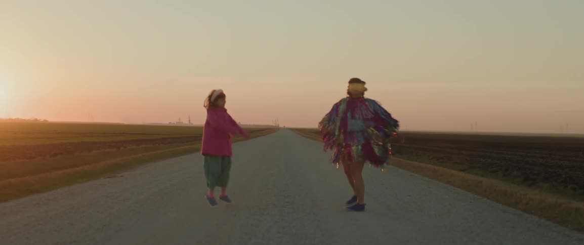 Woman dancing next to a child in the middle of a country road with the sunset on the horizon