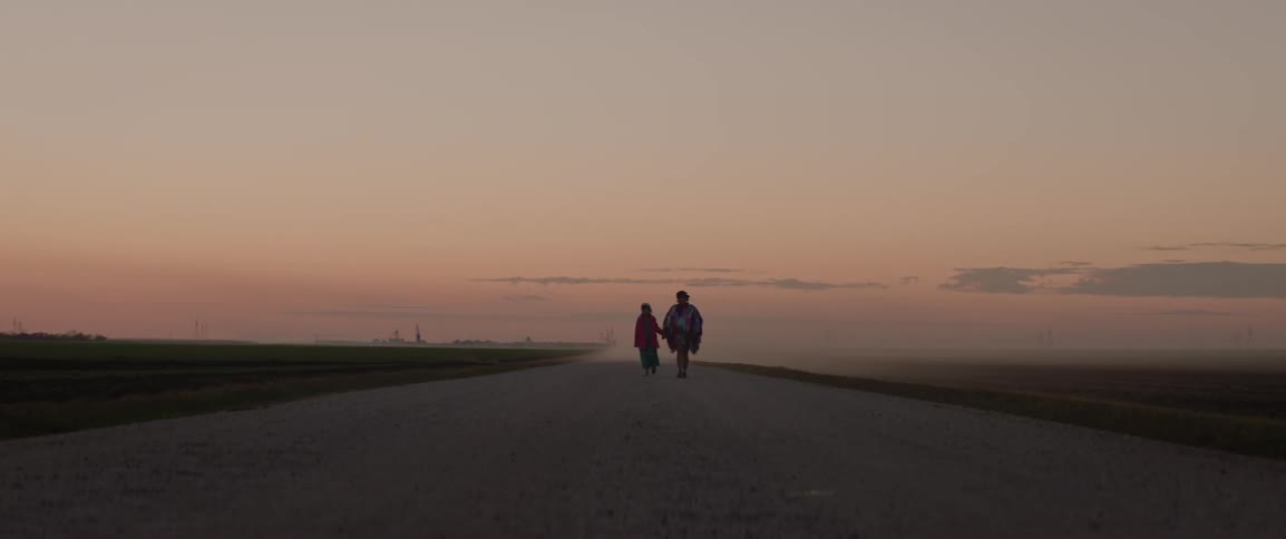 Woman and girl walk hand in hand down farm road passing fields under a red tinted sky