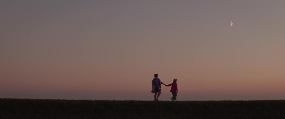 An adult and child walk across the field at dusk under a crescent moon