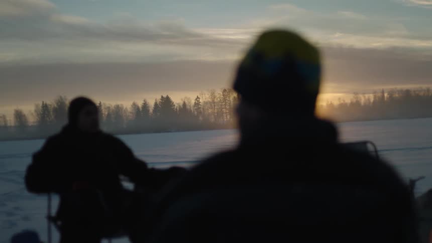 Two people sit in chairs near a campfire at dawn on a frozen lake