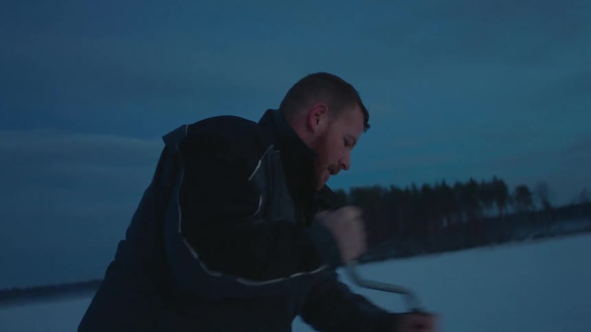 A man uses a hand drill to drill through the surface of a frozen lake at dawn