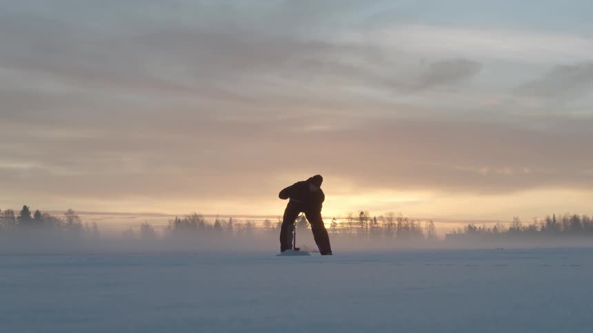 A person drills into a frozen lake at sunrise