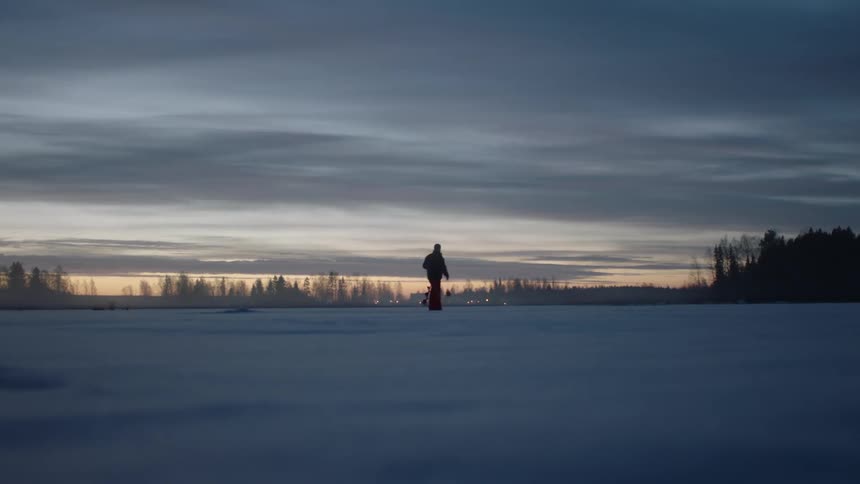 A silhouetted man walks across a snowy field at dusk