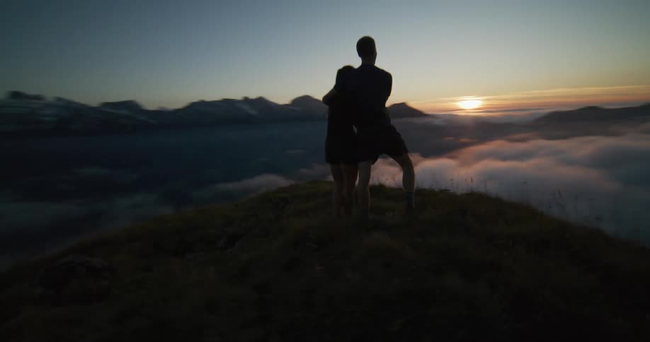 Man and woman stand together on mountain looking across low lying clouds to sun sitting on the red horizon