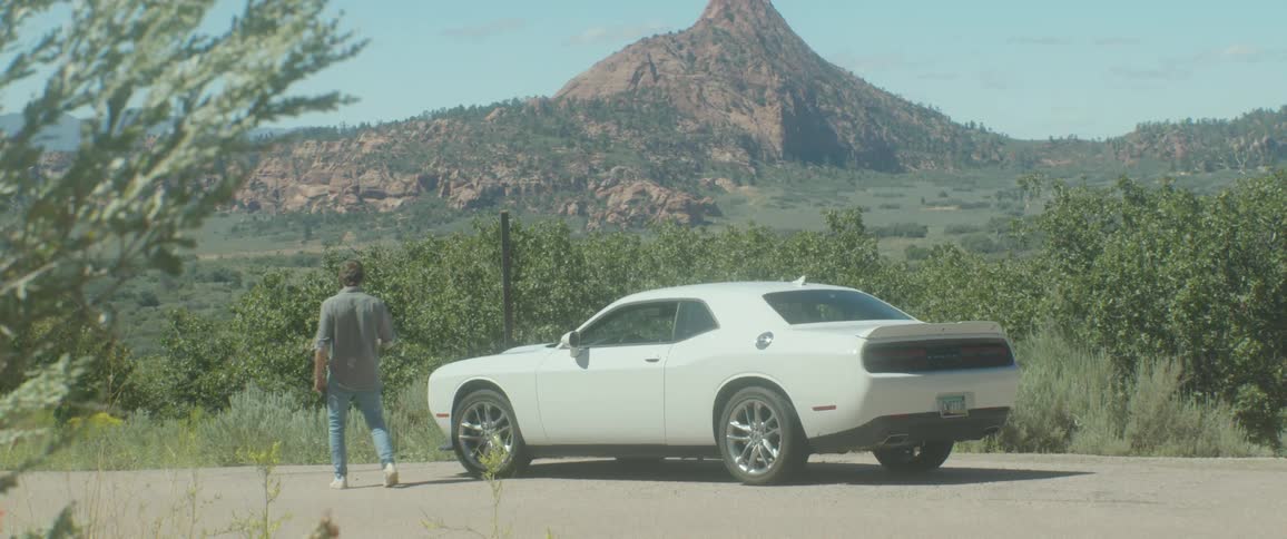 A man walks away from a white dodge charger stopped on the side of a road beside a scenic view of a green landscape with a large mountain in the background on a sunny day
