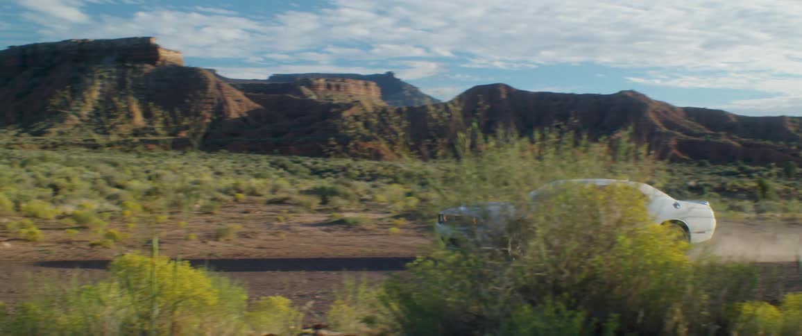 White sports car creates dust while driving past scrub land and mountains on a cloudy day