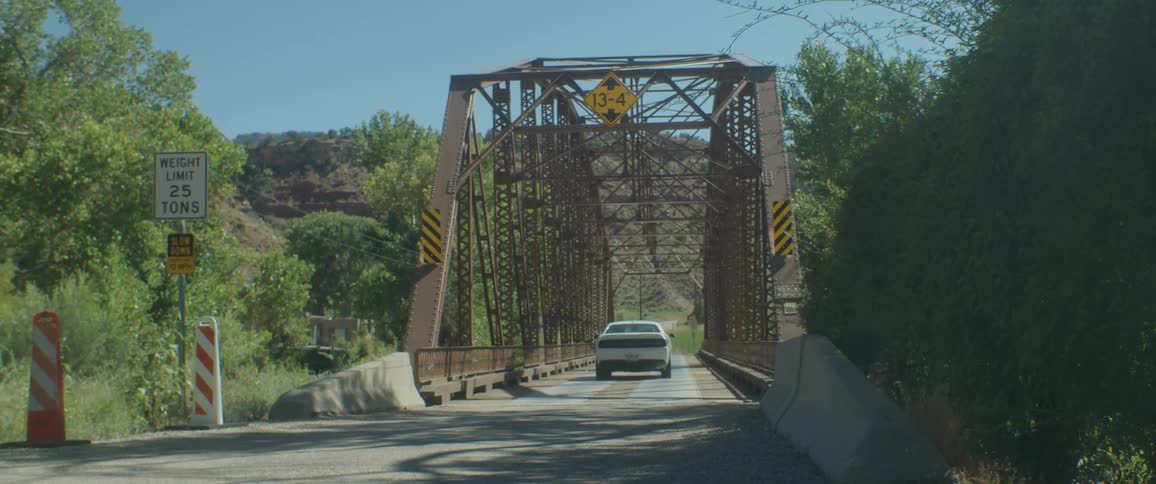 A white dodge car driving by a steel bridge in the rural area