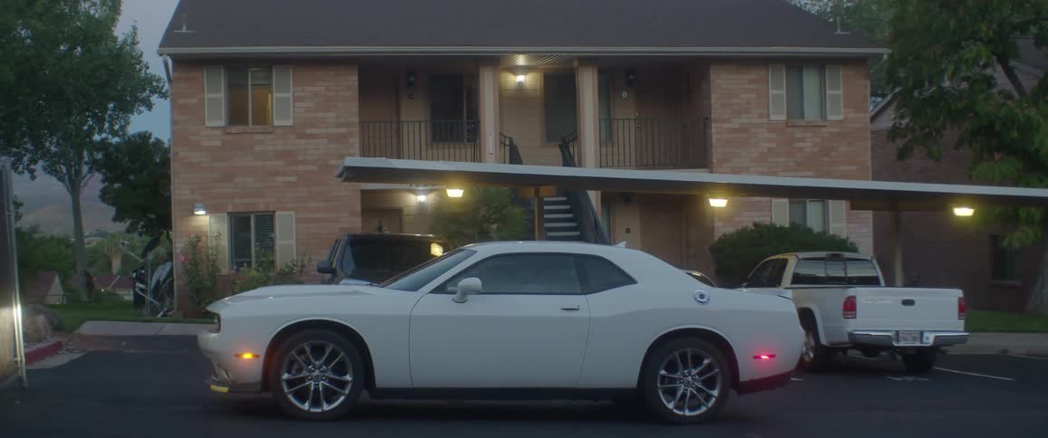 A white car stopping in front of a brick house with other parked cars