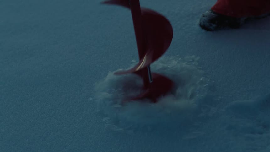 An ice fisherman bores out a fishing hole through the snow and ice