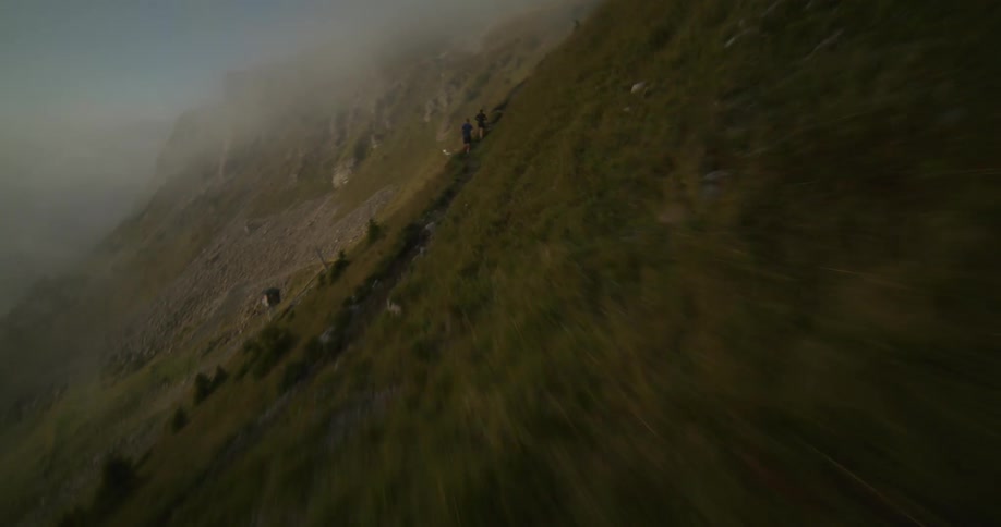 Aerial flyby of a green hillside covered in fog with a trial where two people are jogging