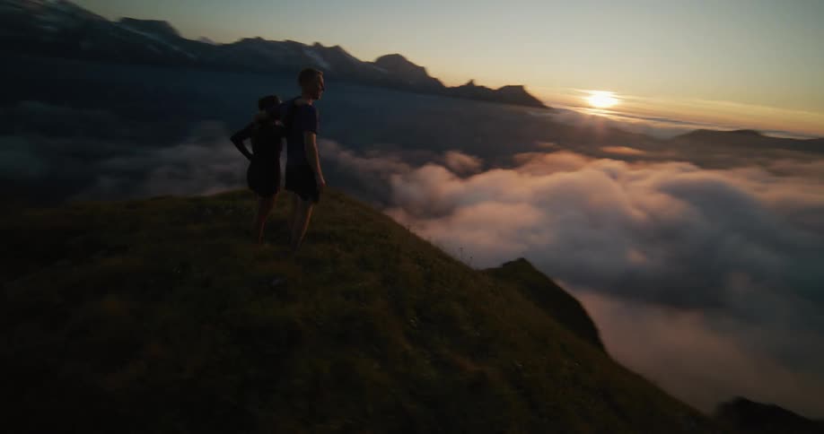 A couple on top of a mountain are holding each other and looking at the horizon with the sun above the clouds
