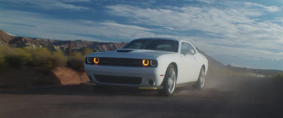 A white sports car slides as it travels fast on a dirt road with mountains in the distance