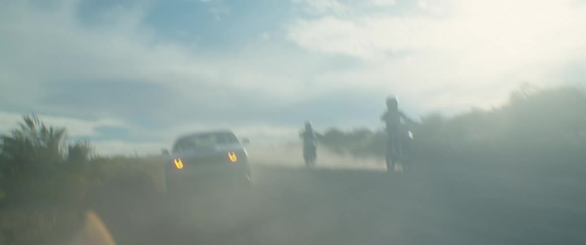 Two motorbikes and a car passing along a dirt road and raising dust
