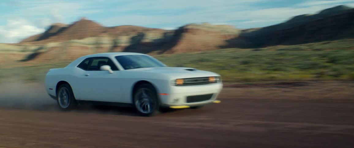 A white car travels fast on a dirt road with mountains in the distance