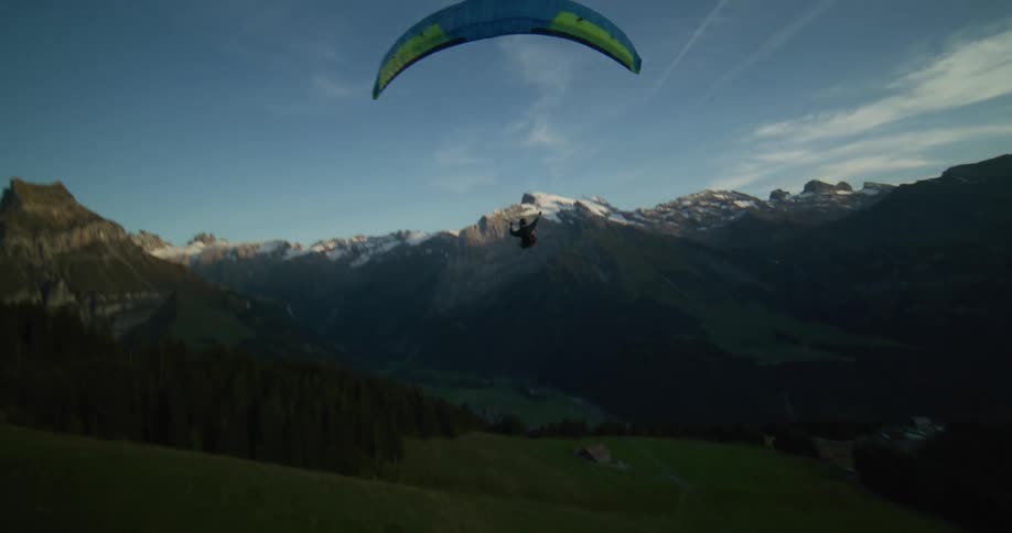 Paraglider flying over a green hillside in the mountains with the sunsetting over white peaks in the distance