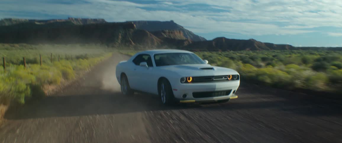 A white sports car slides as it travels fast on a dirt road with mountains in the distance