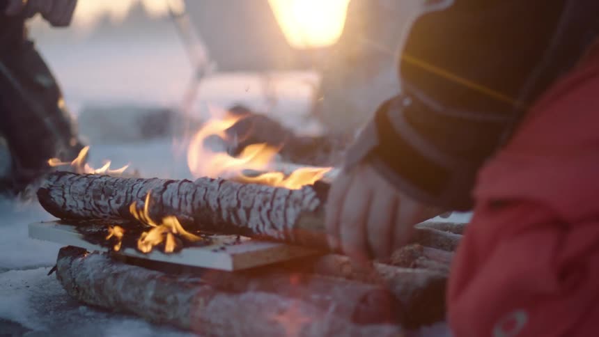 A person adjusts burning logs in the snow at a campsite